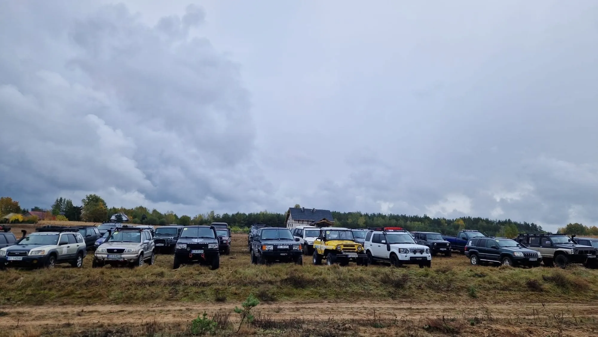 Samochody terenowe uczestników na łące pod szarym, deszczowym niebem, Kulturalny Offroad 2025